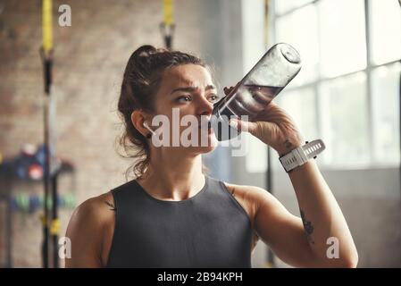 Nahaufnahme der jungen Sportfrau in Ohrhörern ist das Trinkwasser beim Training im Fitnessstudio. Profisport. TRX-Schulung. Gesunder Lebensstil Stockfoto