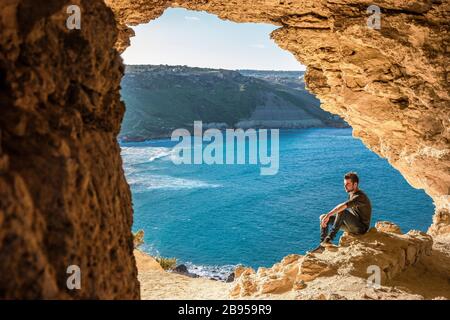 Gozo Island Malta, junger Mann in einer Höhle mit Blick auf den Ozean und Blick auf die Ramla Bay, von innen Tal Mixta Cave Gozo Stockfoto