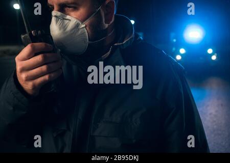 Polizeimann mit chirurgischer Maske spricht mit Walkie-Talkie und Patrouillenwagen mit Sirenen und blauen Lichtern auf den Ausgangssperren in den Straßen während des Zustandes von A. Stockfoto