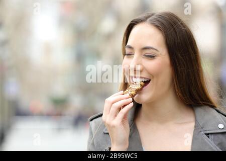 Vorderansicht Porträt eines zufriedenen Mädchens, das eine Getreide-Snack-Bar auf einer Straße in der Stadt isst und genießt Stockfoto