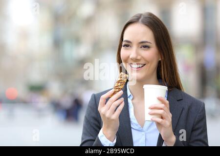 Portrait mit Vorderansicht einer glücklichen Executive-Frau, die eine Getreide-Snack-Bar und einen Kaffee auf der Straße hält Stockfoto