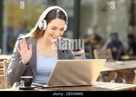 Glückliche Frau, die auf einem Laptop ein schnurloses Headset-Videoband trägt und auf einer Café-Terrasse sitzt Stockfoto