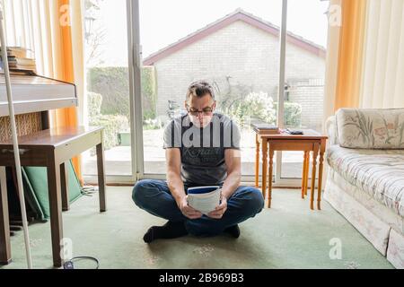 50 etwas männlich in Coronavirus Selbstisolierung zu Hause, sitzen vor dem Fenster im Wohnzimmer und lesen ein Buch Stockfoto