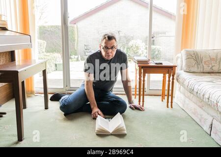 50 etwas männlich in Coronavirus Selbstisolierung zu Hause, sitzen vor dem Fenster im Wohnzimmer und lesen ein Buch Stockfoto