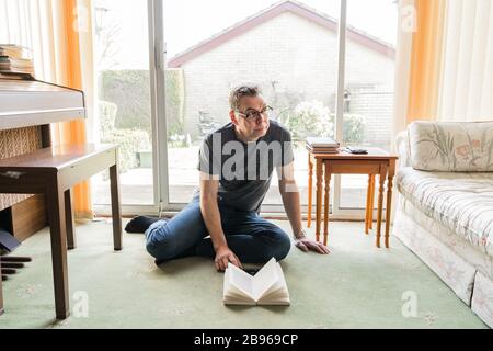 50 etwas männlich in Coronavirus Selbstisolierung zu Hause, sitzen vor dem Fenster im Wohnzimmer und lesen ein Buch Stockfoto