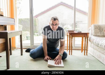 50 etwas männlich in Coronavirus Selbstisolierung zu Hause, sitzen vor dem Fenster im Wohnzimmer und lesen ein Buch Stockfoto