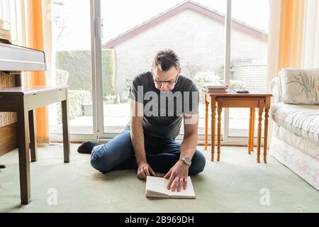 50 etwas männlich in Coronavirus Selbstisolierung zu Hause, sitzen vor dem Fenster im Wohnzimmer und lesen ein Buch Stockfoto