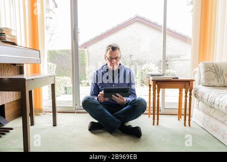 50 etwas männlich in Coronavirus Selbstisolierung zu Hause, sitzend vor dem Fenster im Wohnzimmer, mit Blick auf Tablet-Computer-Pad Stockfoto