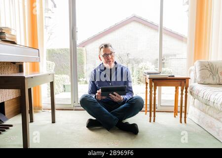 50 etwas männlich in Coronavirus Selbstisolierung zu Hause, sitzend vor dem Fenster im Wohnzimmer, mit Blick auf Tablet-Computer-Pad Stockfoto