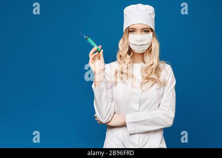 Hübscher weiblicher Arzt mit weißer medizinischer Robe, die Spritze auf blauem Hintergrund hält, isoliert. Konzept für das Gesundheitswesen und die Pharmakologie. Junge Frau in Stockfoto