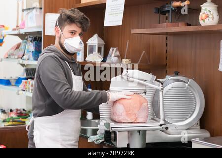 Avetrana, Italien, - Marth 19, 2020. Der Verkäufer serviert einem Kunden einen in Scheiben geschnittenen Schinken, trägt während der Coronavirus Epidemie eine medizinische Maske und Handschuhe. Stockfoto