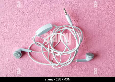 Gebräunte Ohrhörer auf einem texturierten, rosafarbenen Hintergrund. Kopfhörer mit weißen Kabelgewirr. Modernes persönliches Gerät für Musik und Kommunikation. Draufsicht. Stockfoto