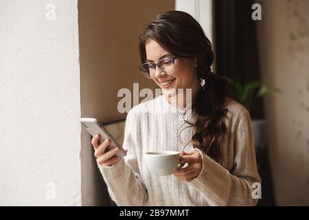 Bild einer jungen, schönen kaukasischen Frau mit langen braunen Haaren, die das Handy hält und Kaffee im Café trinkt Stockfoto