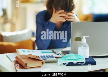 Nahaufnahme von medizinischer Maske und Handdesinfektionsmittel und gestresste Frau im Hintergrund im temporären Heimbüro während der Coronavirus-Epidemie im Haus i Stockfoto