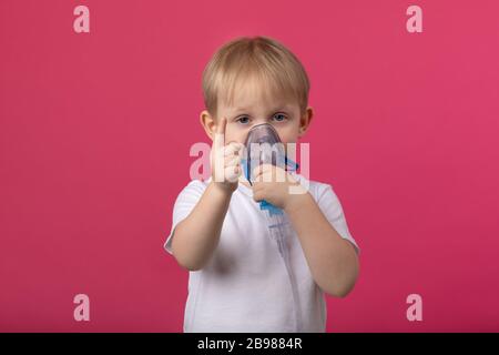 Ein blondes Kind mit einem Inhalator in der Hand, der an seinem Mund befestigt ist, zeigt einen Finger auf einem einfarbig rosafarbenen Hintergrund. Studiofotografie für medizinische Themen von t Stockfoto