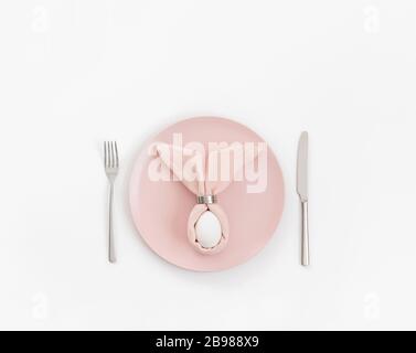 Frohes osterkonzept. Ostertischeinstellung mit Ei in rosafarbener Serviette Osterhase auf weißem Hintergrund. Kopierbereich, Draufsicht, flaches Layout. Stockfoto
