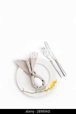 Ostertischeinstellung mit Ei in grauer Serviette Osterhase und einem Zweig gelber Forsythia Blumen auf weißem Grund. Vertikale Ausrichtung. Stockfoto