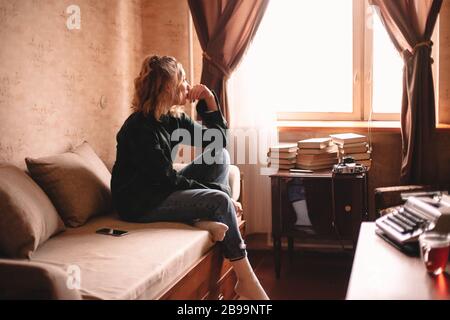 Nachdenkliche junge Frau, die durch Fenster schaut, während sie zu Hause auf dem Bett sitzt Stockfoto