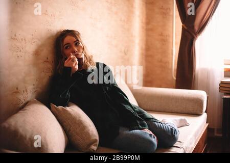 Junge nachdenkliche Frau trinkt Tee, während sie auf dem Sofa im Wohnzimmer zu Hause sitzt Stockfoto