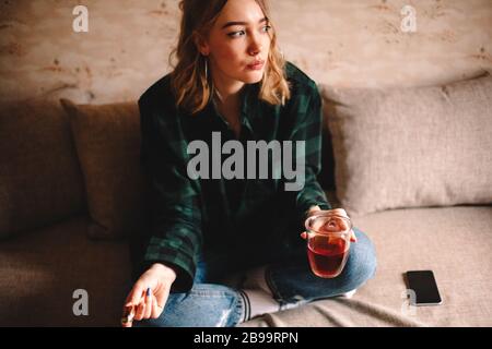 Junge nachdenkliche Frau, die eine Tasse Tee und Schokolade hält, während sie zu Hause auf dem Sofa sitzt Stockfoto