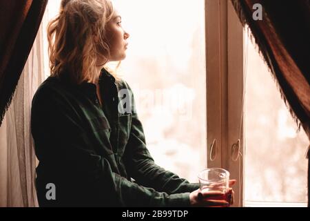 Junge nachdenkliche Frau trinkt Tee und schaut durch Fenster, während sie zu Hause auf Fensterbank sitzt Stockfoto