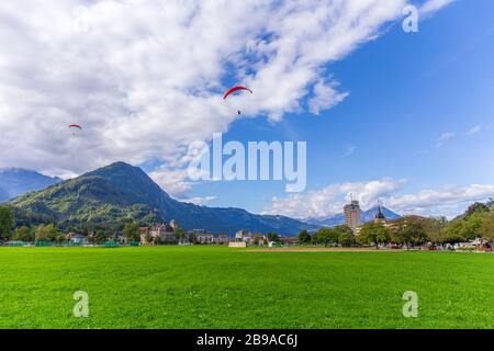 Interlaken, Schweiz - 28.09.2019 - landschaftlich grünes Feld mit Gleitschirm über Bergkette und blauem Himmel. Interlaken liegt zwischen dem Stockfoto