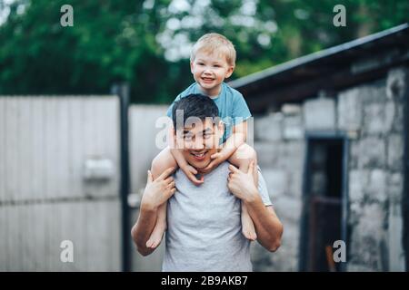 Blonder dreijähriger Junge sitzt auf den Schultern des Vaters. Kasachischer Vater und kaukasische Mutter. Glücklicher Vater mit schwarzen Haaren spielt mit seinem kaukasischen Sohn Stockfoto