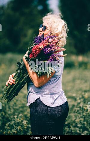 Blonde, reife Stutterin, die einen Lupinenstrauß auf dem Naturhintergrund hält. Das Konzept der Schönheit und des Körpers positiv. Licht bei Sonnenuntergang Stockfoto