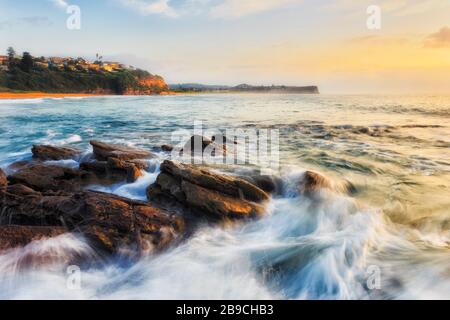 Der hohe Schlüssel der schnellen Wellen, die bei Sonnenaufgang über Sandsteinfelsen am Warriewood Beach in Sydney fließen. Stockfoto