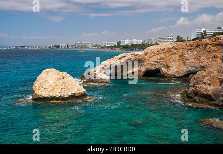 Love Bridge - eine Felsenbrücke natürlichen Ursprungs über das kristallklare Wasser von Kap Greco mit der Ayia Napa Stadt im Hintergrund. Zypern Stockfoto