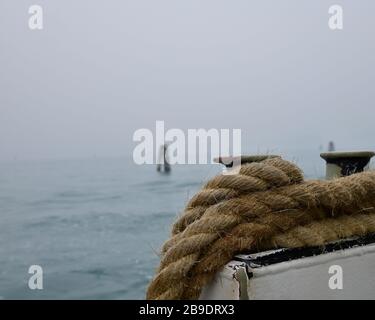 Nahaufnahme des dicken Seils, das um den Schiffssollard auf dem Geländer gebunden ist, mit im Hintergrund der nebligen Lagune von Venedig mit Holz-Anlegepfosten Stockfoto