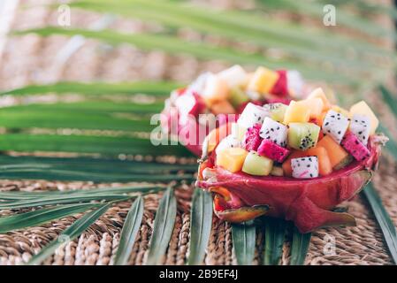 Exotischer Obstsalat in der Hälfte der Drachenfrucht über Palmblättern auf Rattanhintergrund. Kopierbereich. Tropische Reise, exotische Früchte. Veganer und Vegetari Stockfoto