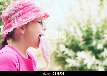 Kleines Mädchen in einem pinkfarbenen Hut leckt großen Lollipop Stockfoto