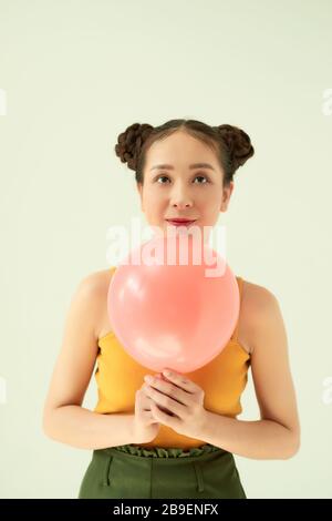 Nettes heiteres asiatisches Teenager-Mädchen, das einen rosafarbenen Luftballon mit zwei Brötchen Frisur über hellem Hintergrund hält. Stockfoto