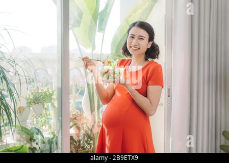 Voller Vitamine. Energisch gut aussehende schwangere Frau, die ihr Fleisch isst, während sie Teller in der Hand trägt und sich am Fenster entspannt Stockfoto
