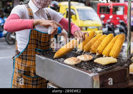Der Koch streut Salz auf frisch gegrilltem Mais, Straßennahrung in Asien, Thailand. Stockfoto