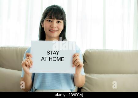 Asian Woman, die ein Blatt Papier mit Text hält, bleibt zu Hause, um Virus zu schützen und kümmert sich um ihre Gesundheit von COVID-19. Bleiben Sie zu Hause. Stockfoto