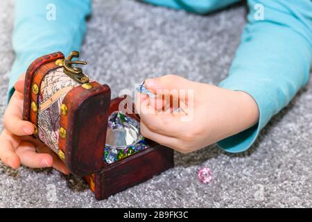 Fünf Jahre alter Junge, der mit einer Schatzkiste voller bunter Juwelen spielt. Der Junge hat ein türkisfarbenes Hemd und hält ein blaues Schmuckstück. Stockfoto