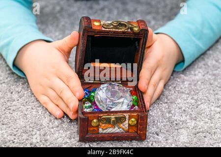 Fünf Jahre alter Junge, der eine Schatzkiste voller bunter Juwelen in den Händen hält. Der Junge hat ein türkisfarbenes Hemd. Stockfoto