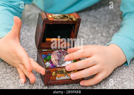 Fünf Jahre alter Junge, der mit einer Schatzkiste voller bunter Juwelen spielt. Der Junge hat ein türkisfarbenes Hemd. Stockfoto