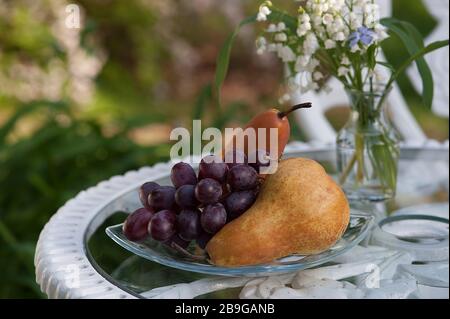 Trauben und Birnen auf einer Glasschale draußen auf einem weißen schmiedeeisernen Tisch in einer Gartenanlage. Stockfoto