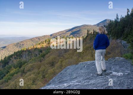 Frau, die Fleece-Jacke trägt und vom Felsvorsprung auf dem Long Trail aus den Blick auf die Berge hat, auf dem Weg zum Mt. Grant in Lincoln, Vermont Stockfoto