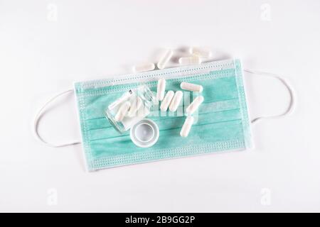 Weiße Arzneikapseln, die aus der Glasflasche auf einer chirurgischen Maske verschüttet werden (Draufsicht auf weißem Hintergrund) Stockfoto
