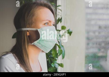 Frau in medizinischer Maske in der Nähe des Fensters. Quarantäne zu Hause. Stockfoto