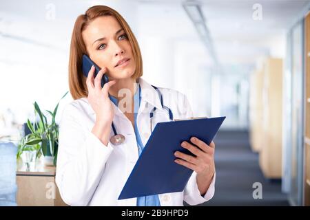Schuss von Frauen mittleren Alters, die mit ihrem Patienten auf dem Handy sprechen, während sie in der medizinischen Klinik stehen. Stockfoto