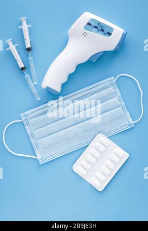 Pandemie des Coronavirus (COVID-19). Medizinische Schutzmaske, Pillen, elektronisches Infrarot-Thermometer und zwei Spritzen auf blauem Hintergrund. Konzept von med Stockfoto