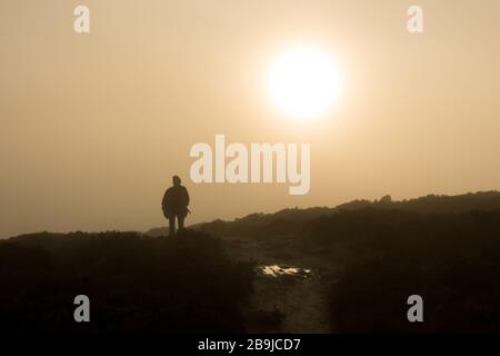 Stimmungsvoller Blick auf eine Person, die in einer Wolkeninversion auf dem Land, Ilkley Moor, West Yorkshire, Großbritannien, aus einem nebligen Tal in den Sonnenschein spazieren geht Stockfoto