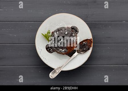 Ein einzelner Schokoladenschwammkuchen mit einer klebrigen Schokoladensoße auf einem weißen Teller mit einem Löffel, auf einer dunkelgrauen Holzoberfläche mit Platz für c geschossen Stockfoto