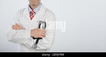 Stethoskop in den Händen eines bärtigen Arztes mit gekreuzten Armen. Arzt in einem blauen Hemd, roter Krawatte und weißem medizinischen Mantel Stockfoto