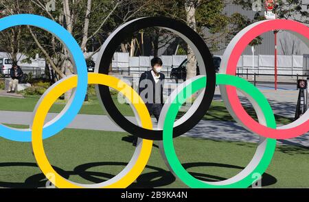 Tokio. März 2020. Das Dateifoto vom 12. März 2020 zeigt einen Fußgänger, der eine Maske trägt, die an den olympischen Ringen in Tokio, Japan vorbeiläuft. Der japanische Premierminister Shinzo Abe teilte am Dienstag mit, dass Japan und das Internationale Olympische Komitee (IOC) vereinbart haben, die Olympischen und Paralympischen Spiele in Tokio um ein Jahr zu verschieben. Credit: Du Xiaoyi/Xinhua/Alamy Live News Stockfoto